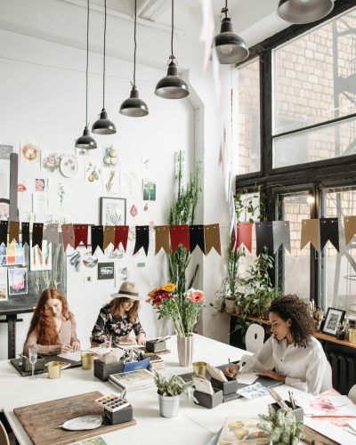 Women engaged in artistic activities at a lively indoor workshop filled with decorations and natural light.